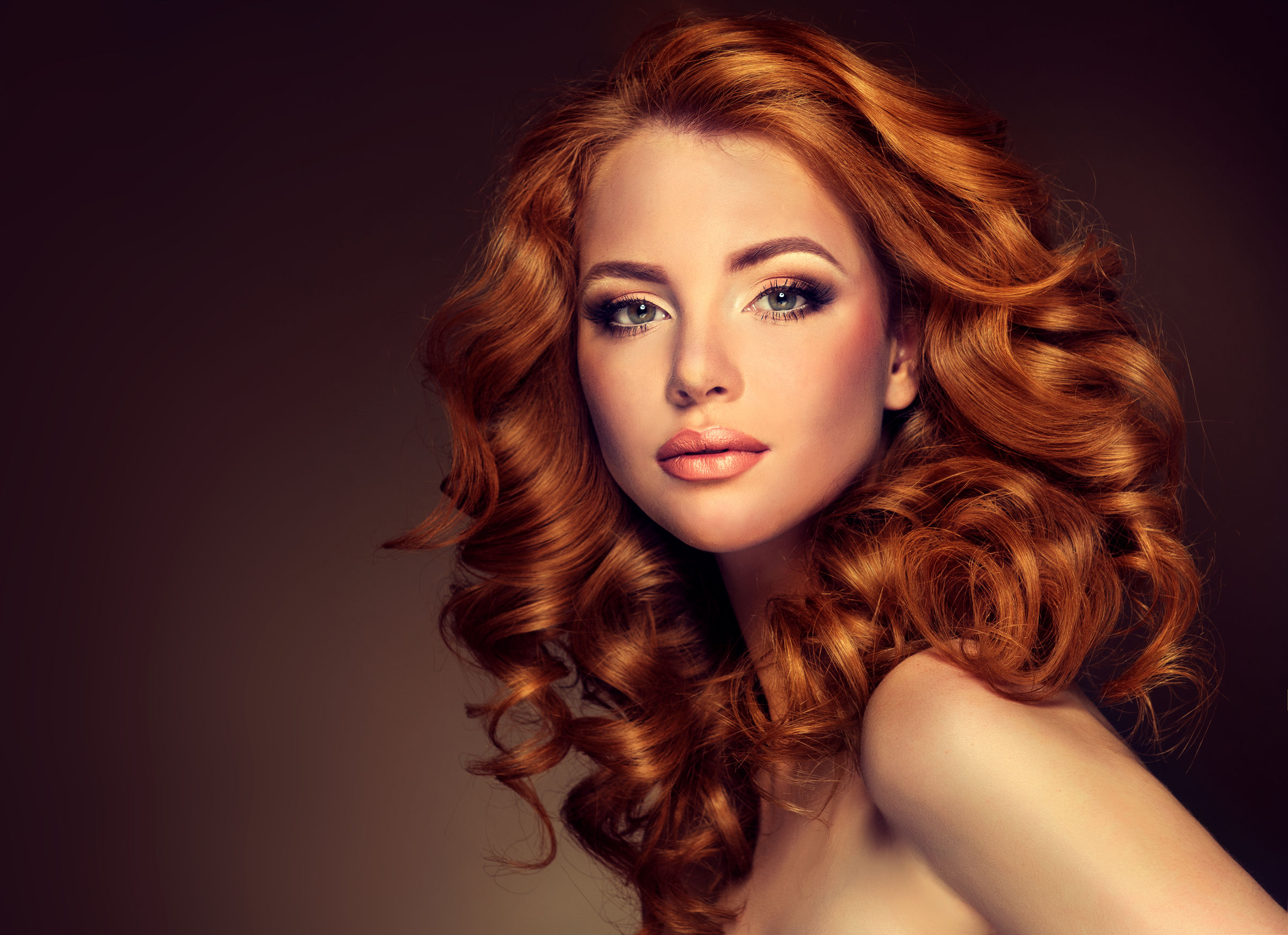 Splash Hair Salon A striking portrait of a woman with vibrant, voluminous red curls cascading around her face and shoulders. Her makeup is elegantly applied, accentuating her eyes and lips, while the warm background complements her hair color, creating a glamorous and sophisticated look.
